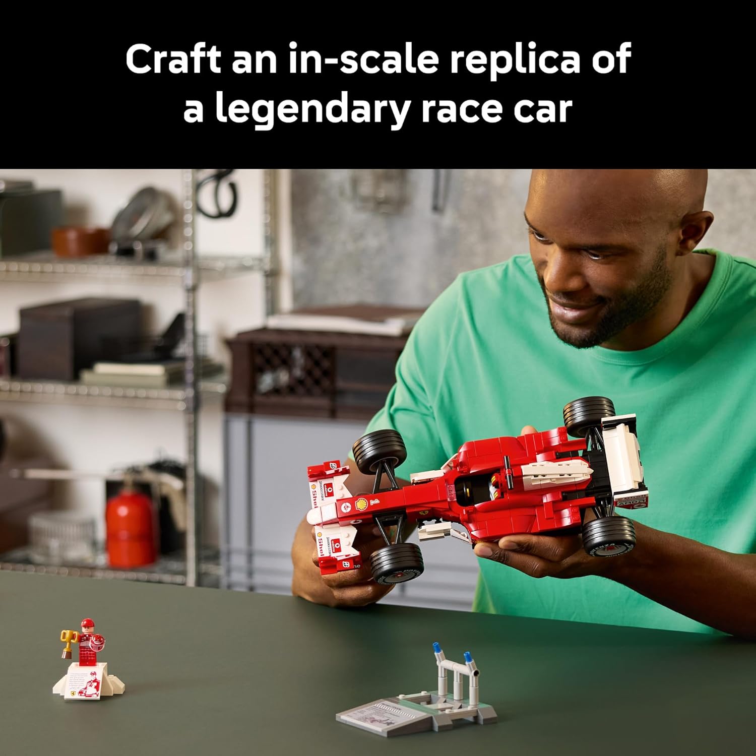 Man holding a red toy race car with small figures on a table in a workshop setting.